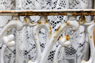 Rusty white balcony railing with decorative floral design and lace curtain in background