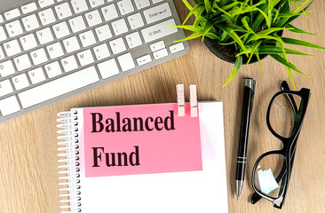 Balanced fund phrase on pink note attached to spiral notebook with keyboard, pen, glasses and plant
