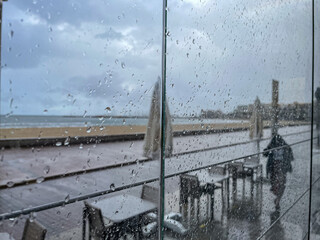 Moody rainy scene viewed through window with raindrops, featuring empty outdoor café tables and a...