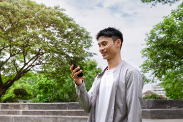 A young man stands playing with his phone in a park.