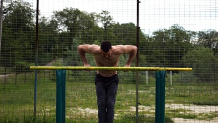 Hardy shirtless sportsman doing exercises on parallel bars at nature. Strong and muscular man working out at sports ground. Young athletic guy exercising outdoor. Concept of active healthy lifestyle
