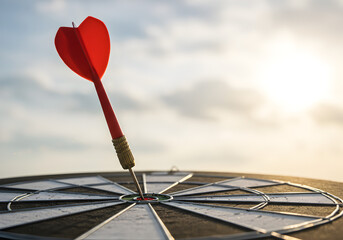 Red dart precisely hitting the bullseye of a dartboard under a bright, cloud-dotted sky, symbolizing perfect aim and goal achievement.