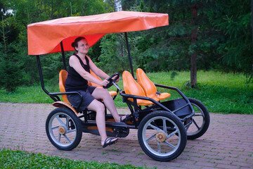Woman riding a four-wheeled pedal bike on a paved path in a green park. Outdoor recreation and...