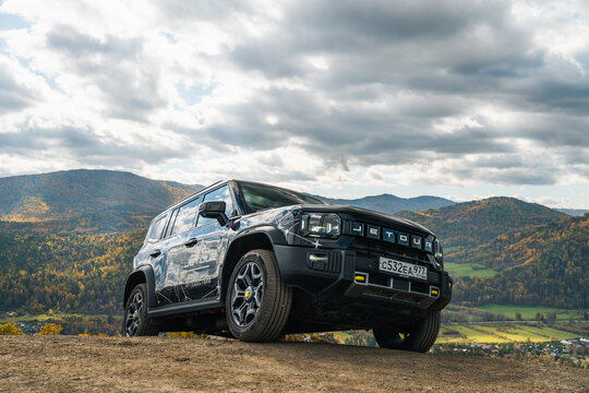 Aktash, Altai Republic - October 12, 2023: A black Jetour T2 SUV is parked on a grassy hill with a scenic mountain range covered in autumn trees under a cloudy sky in the background
