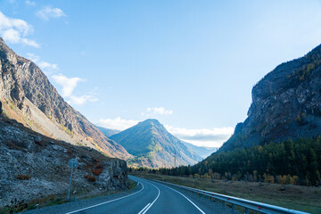 Naklejka premium A wide, panoramic view of a winding road leading into a valley with mountains covered in autumn trees under a bright blue sky