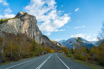 A winding road leads into the distance with a tall, rocky mountain on the left side and a forest of autumn trees on the right