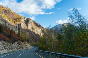 A view of a winding asphalt road with a guardrail, surrounded by a rocky mountainside and a forest...