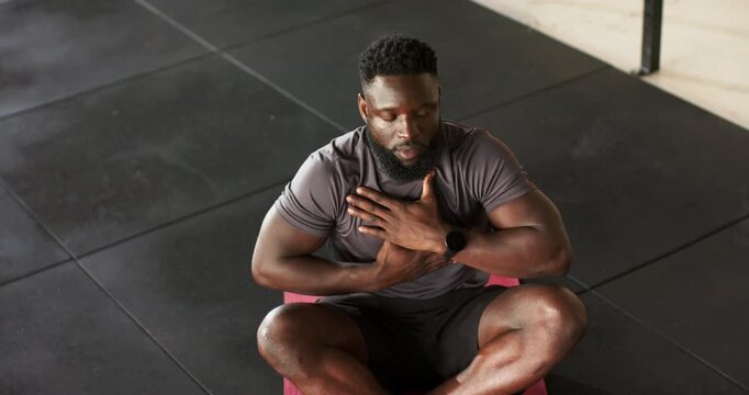 Settling cross-legged on pink mat, African American man breathing for focus at gym with smartwatch