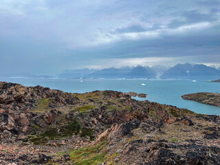 A scenic view from Scoresby Land in Greenland with rocks, water, icebergs, and mountains on an...