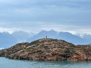An unidentifiable person standing on the top of a rocky hill in Scoresby Land in Greenland with...