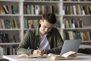 Productive self studying. Smiling diligent male high school student write creative academic work prepare for test exam conduct scientific research reviewing printed books digital literature via laptop