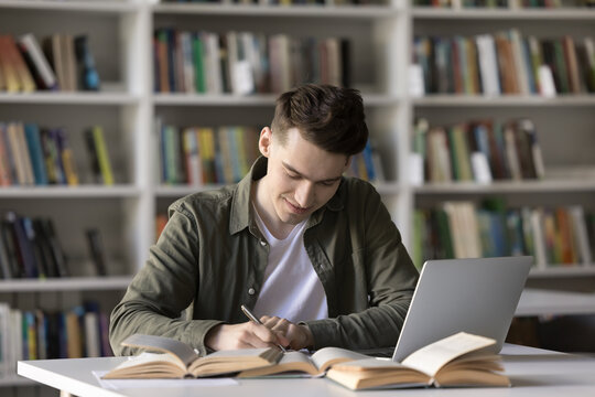 Productive self studying. Smiling diligent male high school student write creative academic work prepare for test exam conduct scientific research reviewing printed books digital literature via laptop