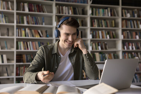 Hybrid learning tools. Smart smiling gen z male high school student sit in earphones before notebook look at screen attend virtual lecture webinar video call consultation prepare to take paper notes