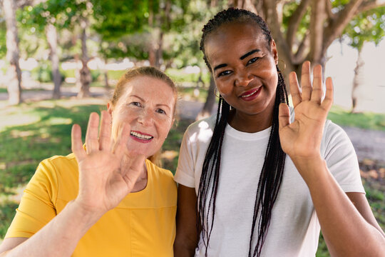 Diverse female friends smiling, waving to the camera together during sunny outdoor meetup near lush greenery with trees surrounding them - Powered by Adobe