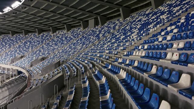 Aerial view drone flies over empty stadium or race track seats. Rows and blue and white seats without viewers and spectators. Evening shot of empty football stadium. Soccer Arena drone aerial view.
