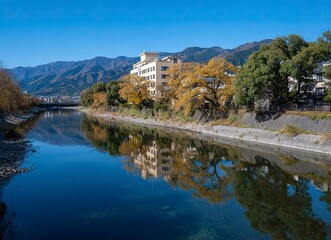 Fototapeta premium The riverbank of the Ohtawara River in Nagano, Japan is a fantastic photograph, with a clear sky and reflection on the water surface, a large building across the street, and trees along the embankment