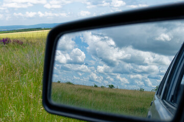The car mirror reflects a bright, cloudy sky and a vast, grassy field on a sunny day.