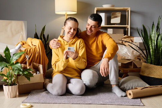 Happy young couple in a new home, sitting on the floor surrounded by moving boxes, looking at a phone and laughing.