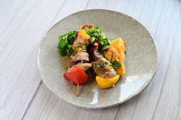 Top-down view of a colorful plated meal featuring a grilled chicken skewer, fresh salad, and creamy polenta on a white background.