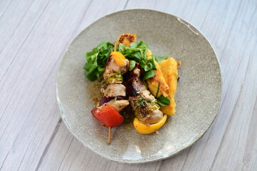 Top-down view of a colorful plated meal featuring a grilled chicken skewer, fresh salad, and creamy polenta on a white background.
