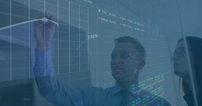 Colleagues wearing business attire annotating financial charts on office glass board, using marker