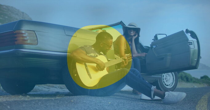 Man strumming acoustic guitar on coastal road near silver convertible, with sun hat and sunglasses - Powered by Adobe