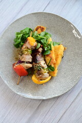 Top-down view of a colorful plated meal featuring a grilled chicken skewer, fresh salad, and creamy polenta on a white background.
