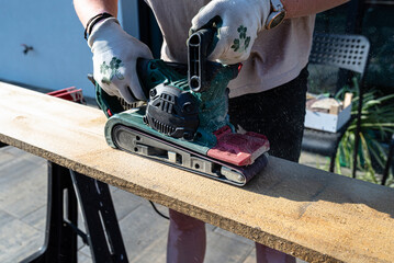 A woman grinds a board with a belt sander on trestles in the garden.