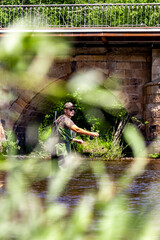 Fisherman casting in river near stone bridge, framed by greenery