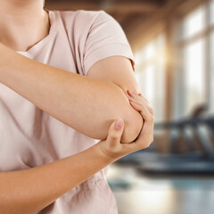 Elbow pain and injury – hands holding painful arm, fitness area background. Sport workout,...
