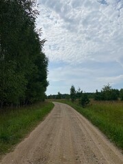 road in the countryside Rural unpaved road through green field with trees under cloudy sky. Nature landscape path for journey and travel.