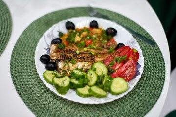 Grilled fish with vibrant vegetables and olives served on white plate. Decorative green mat adds texture. Bright natural light enhances fresh ingredients colors