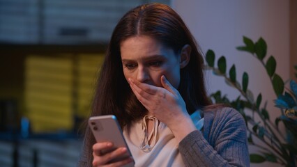 Caucasian woman holding mobile phone read message shock negative bad news at home night evening...
