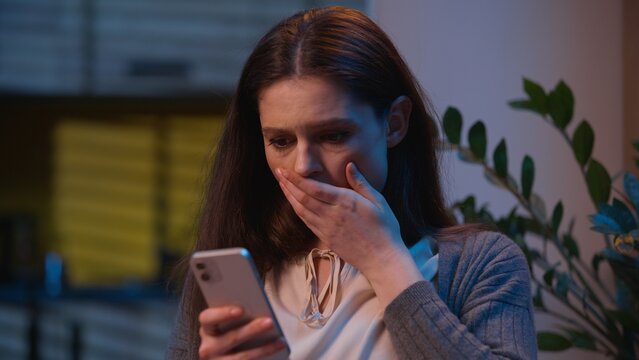 Caucasian woman holding mobile phone read message shock negative bad news at home night evening apartment upset female 30s girl wife shocked failure scam afraid internet smartphone bank reject problem