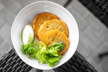 Golden potato pancakes served with sour cream and herb oil, garnished with fresh greens in a white bowl on a rustic woven surface, top view of appetizing vegetarian dish.