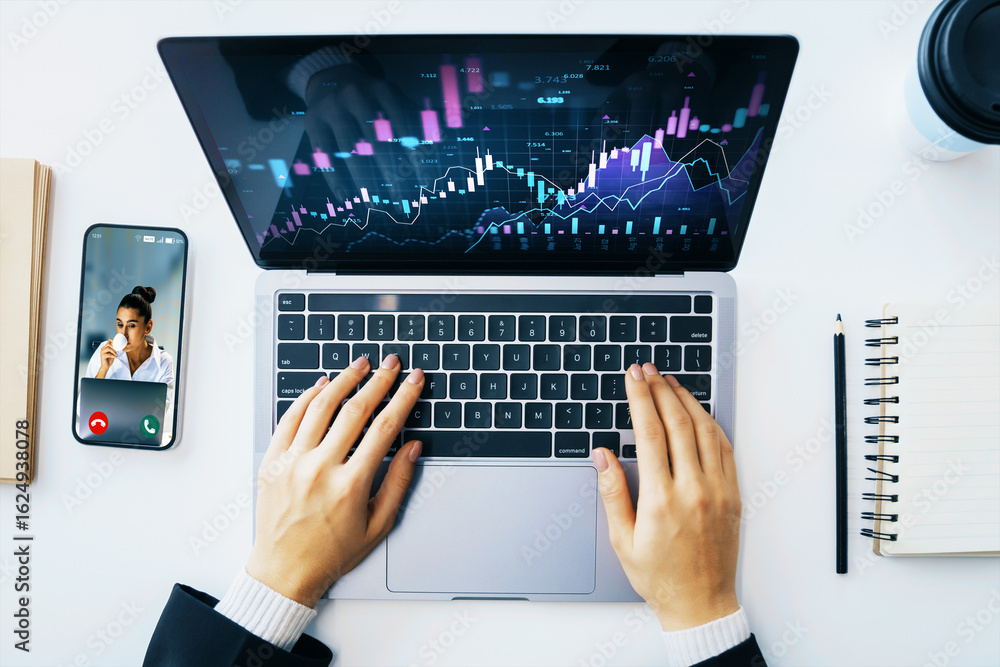 Poster trader working on laptop with financial charts on screen and video call on smartphone, top view on w