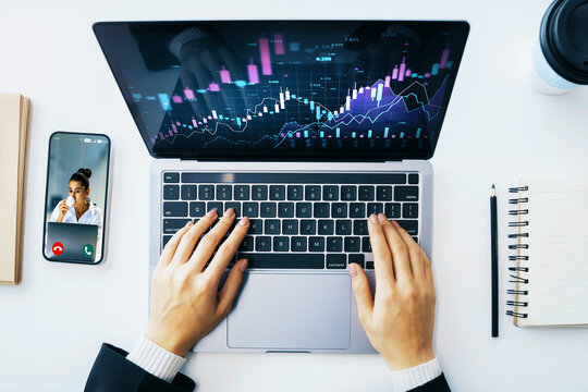 Trader working on laptop with financial charts on screen and video call on smartphone, top view on white office desk with notebook and coffee cup. - Powered by Adobe