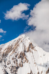 A vertical close-up of a majestic snowcapped mountain peak with rugged, rocky slopes under a bright...