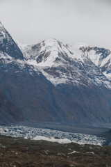 A high-altitude mountain landscape with snowcapped peaks towering over a rugged rocky glacier under a cloudy, overcast sky