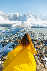 A young Caucasian woman with long brown hair, seen from behind, stands on a rocky ridge in a yellow...
