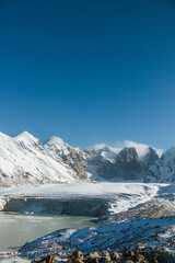 A vertical view of a frozen lake and a glacier in a vast valley with a majestic snowcapped mountain range in the background under a clear blue sky