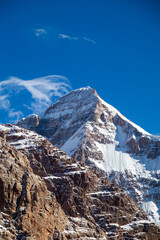 A dramatic vertical close-up of a rugged, snowcapped mountain peak with jagged, rocky slopes under...
