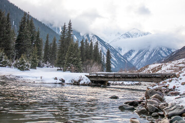 Fototapeta premium A river flows through a snow-covered mountain valley with a small wooden bridge, surrounded by trees and mountains under a cloudy sky