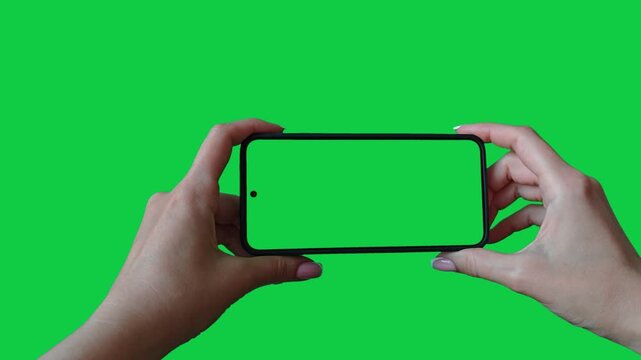 Hands holding cell phone with green screen in green background chroma key. Phone in female hands. Watching video on phone in horizontal position.