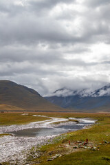 A river winds through a vast, golden and green valley with mountains and low clouds in the...