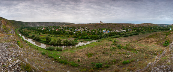 A wide-angle panoramic view of a river valley with a village and a distant monastery under a...