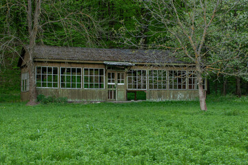 A rustic, long wooden building with large windows and a worn facade, nestled in a lush green field,...