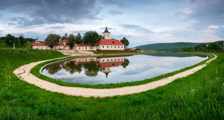 Obraz premium A tranquil panoramic view of a monastery with red-tiled roofs and a bell tower, beautifully reflected in a still pond, representing religious architecture and serene landscapes for travel and spiritua