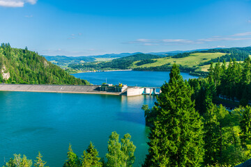 A wide dam structure on a blue lake with forested slopes and rolling fields in the background....