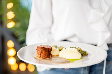 Plate with roasted meat, mashed potatoes, and vegetables in a warm, cozy restaurant setting, held by a casually dressed person